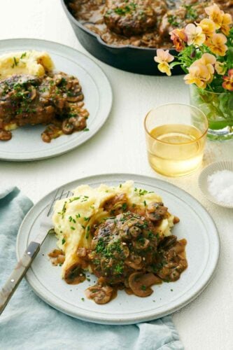 A plate with salisbury steak and mashed potatoes. A second plated serving in the back along with a drink, vase of flower, and skillet.