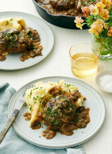 A plate with salisbury steak and mashed potatoes. A second plated serving in the back along with a drink, vase of flower, and skillet.