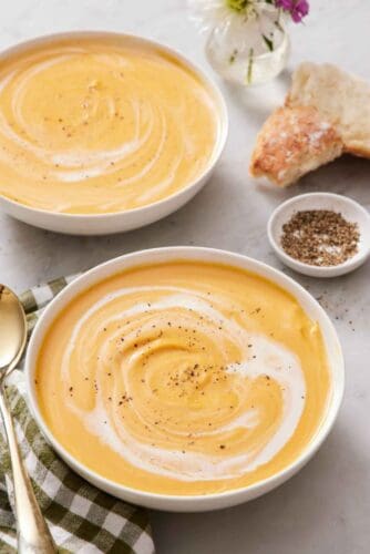 Two bowls of sweet potato soup with cream swirled in and topped with pepper. A bowl of pepper and torn bread on the side.