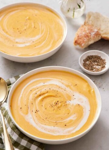 Two bowls of sweet potato soup with cream swirled in and topped with pepper. A bowl of pepper and torn bread on the side.