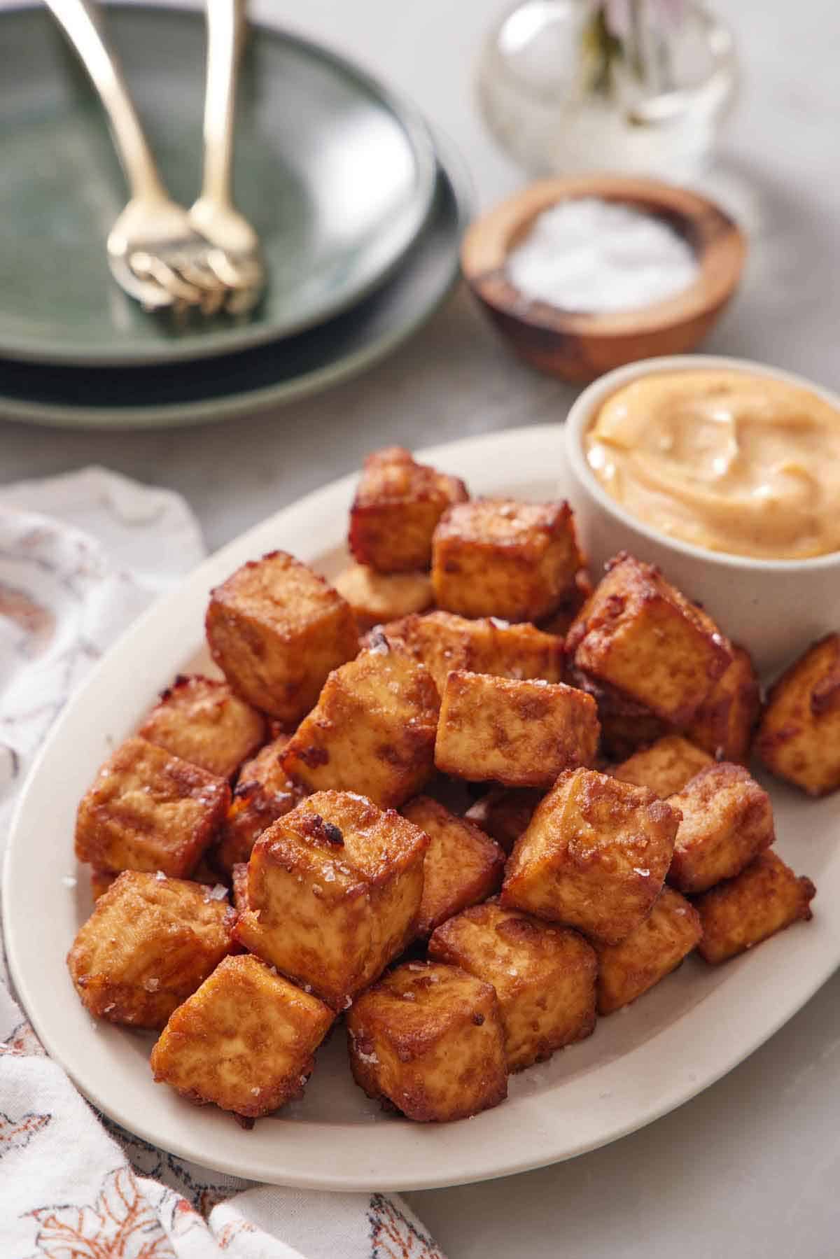 A platter of crispy air fryer tofu with a bowl of dip. A stack of plates, forks, and bowl of salt in the back.