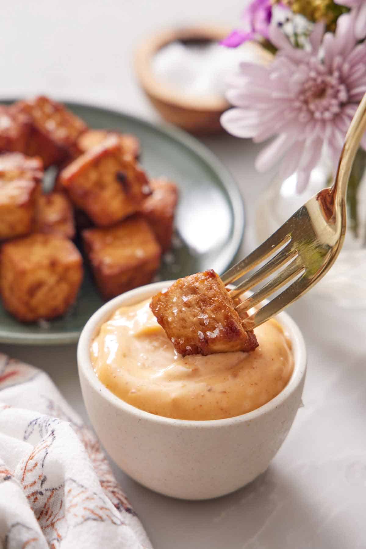 A fork with an air fryer tofu cube dipped into a small bowl of sauce. More tofu in the background.