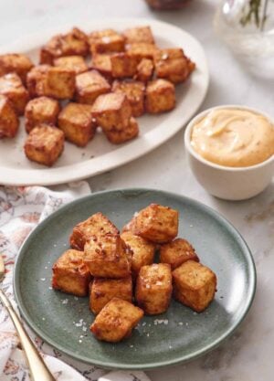 A plate with a pile of air fryer tofu with a platter in the background with more tofu. A bowl of sauce on the side.