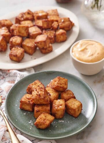 A plate with a pile of air fryer tofu with a platter in the background with more tofu. A bowl of sauce on the side.