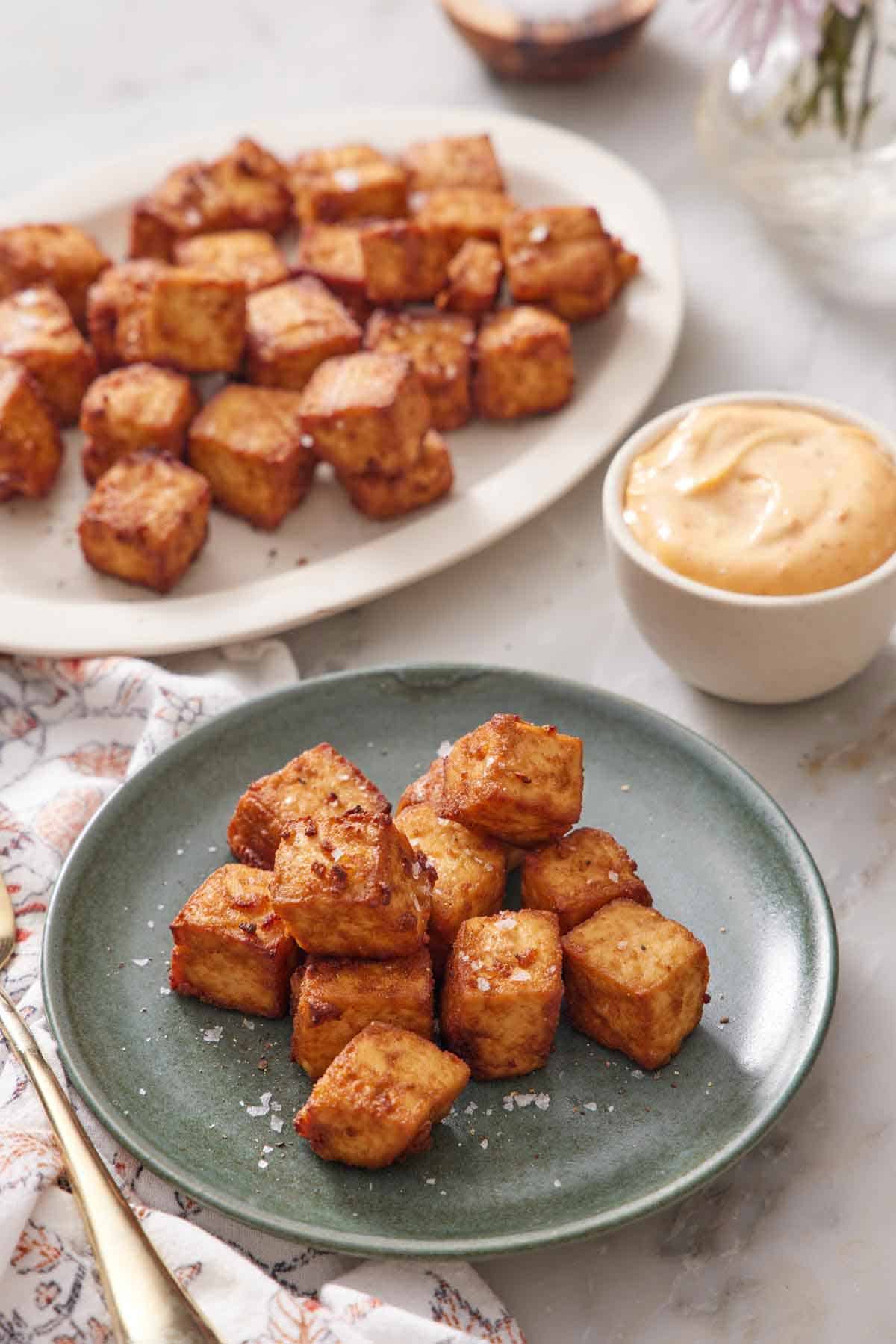 A plate with a pile of air fryer tofu with a platter in the background with more tofu. A bowl of sauce on the side.