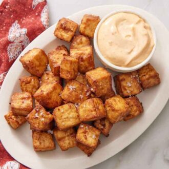 Overhead view of an oval platter of crispy air fryer tofu with a bowl of dip. A linen napkin beside it.