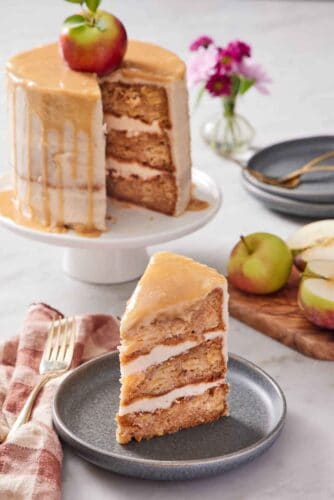 A slice of caramel apple cake on a plate with the rest of the cake in the background on a white cake stand. Apples on the side and some flowers in the back.