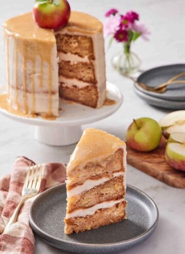 A slice of caramel apple cake on a plate with the rest of the cake in the background on a white cake stand. Apples on the side and some flowers in the back.