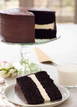 A slice of Ding Dong cake on a plate with a glass of milk and rest of the cake on a cake stand in the background.
