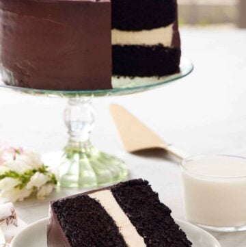 A slice of Ding Dong cake on a plate with a glass of milk and rest of the cake on a cake stand in the background.