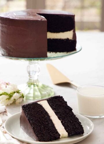 A slice of Ding Dong cake on a plate with a glass of milk and rest of the cake on a cake stand in the background.