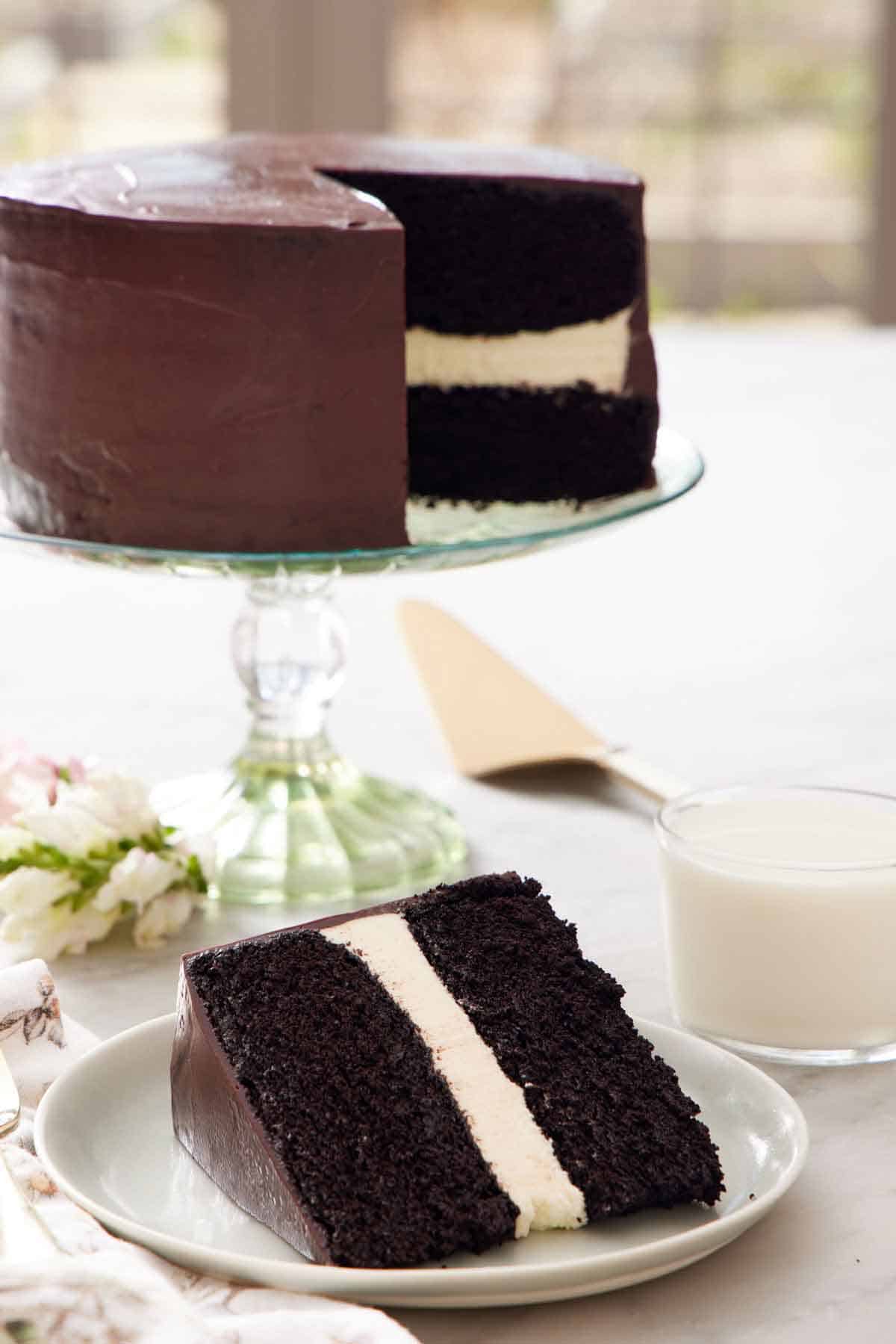 A slice of Ding Dong cake on a plate with a glass of milk and rest of the cake on a cake stand in the background.