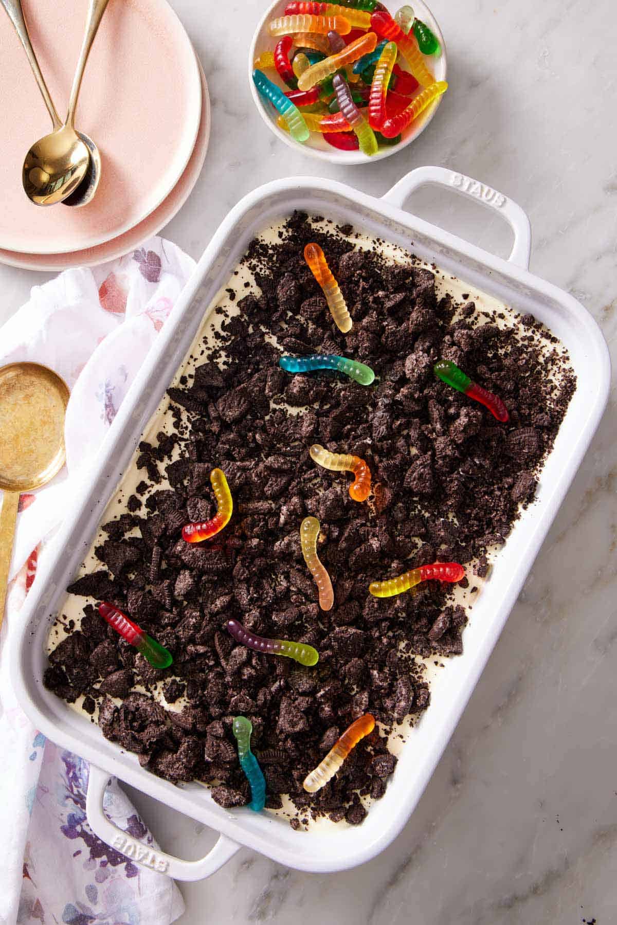 Overhead view of dirt cake in a white baking dish. Gummy bears off to the side in a bowl with some plates and spoons.