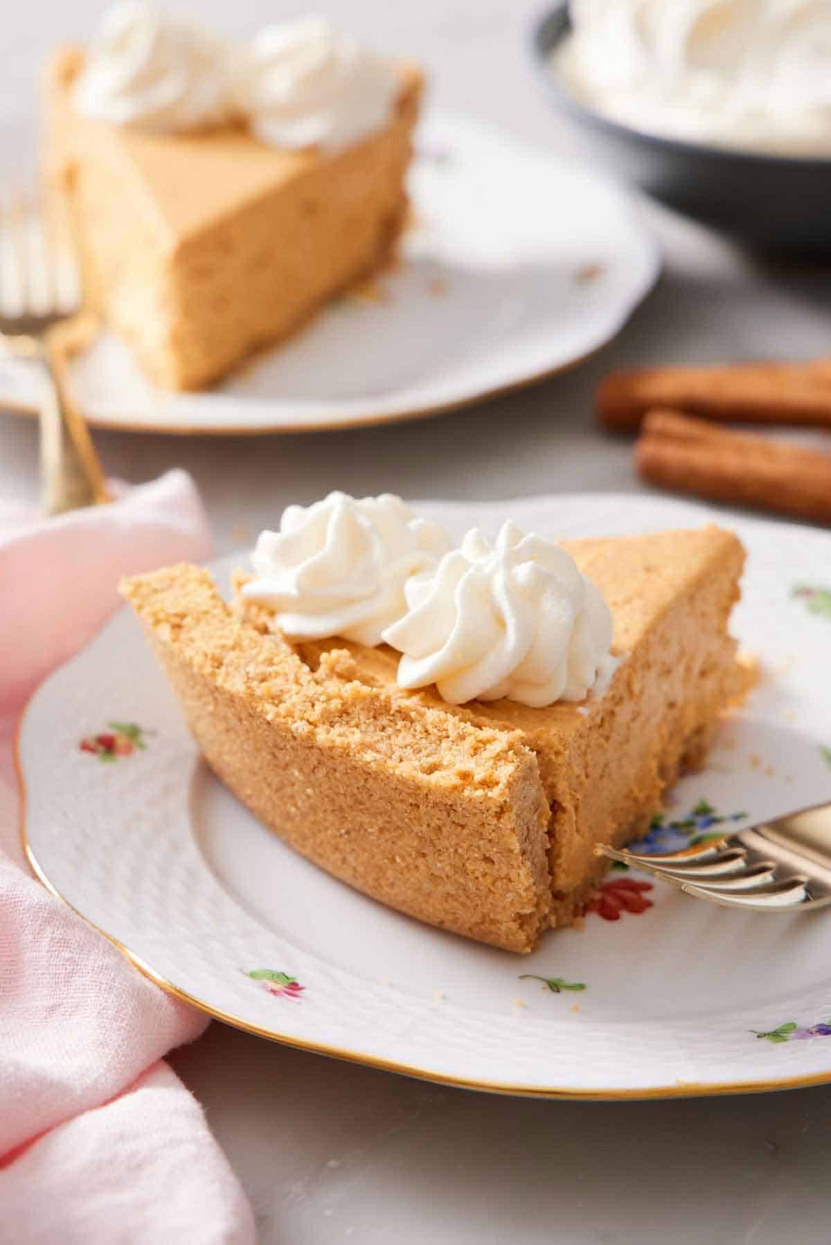 A slice of cheesecake with a graham cracker crust and two dollops of whipped cream on top on a plate with a fork. A second slice in the background.