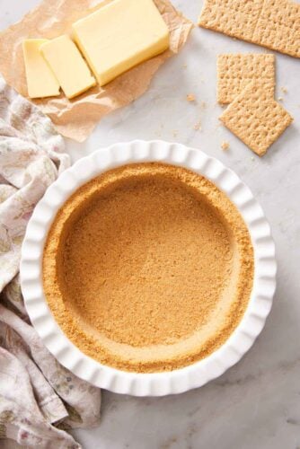 Overhead view of a pie dish containing a graham cracker crust. Butter and graham crackers in the background.