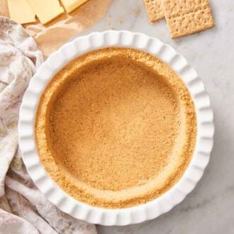 Overhead view of a graham cracker crust in a baking dish. A napkin off to the side with butter and graham crackers.