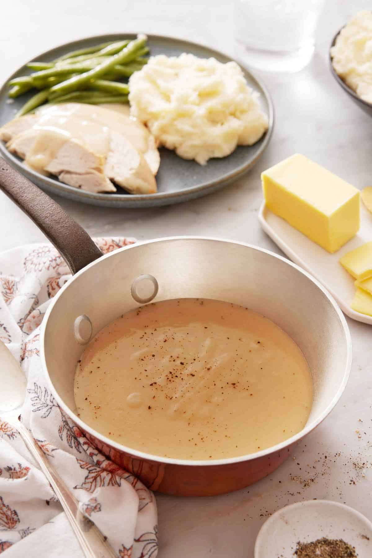 A saucepan of gravy topped pepper. A plate in the background with sliced turkey, mashed potatoes, and green beans. Butter on a separate plate.