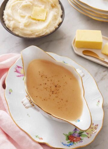 Overhead view of gravy in a gravy boat on a plate. Mashed potatoes in the background with a plate of butter.