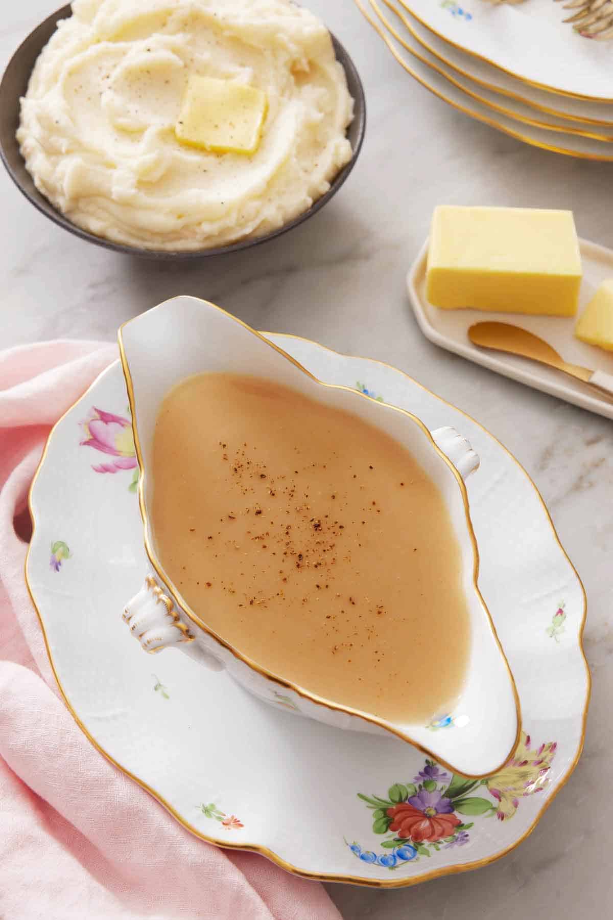 Overhead view of gravy in a gravy boat on a plate. Mashed potatoes in the background with a plate of butter.