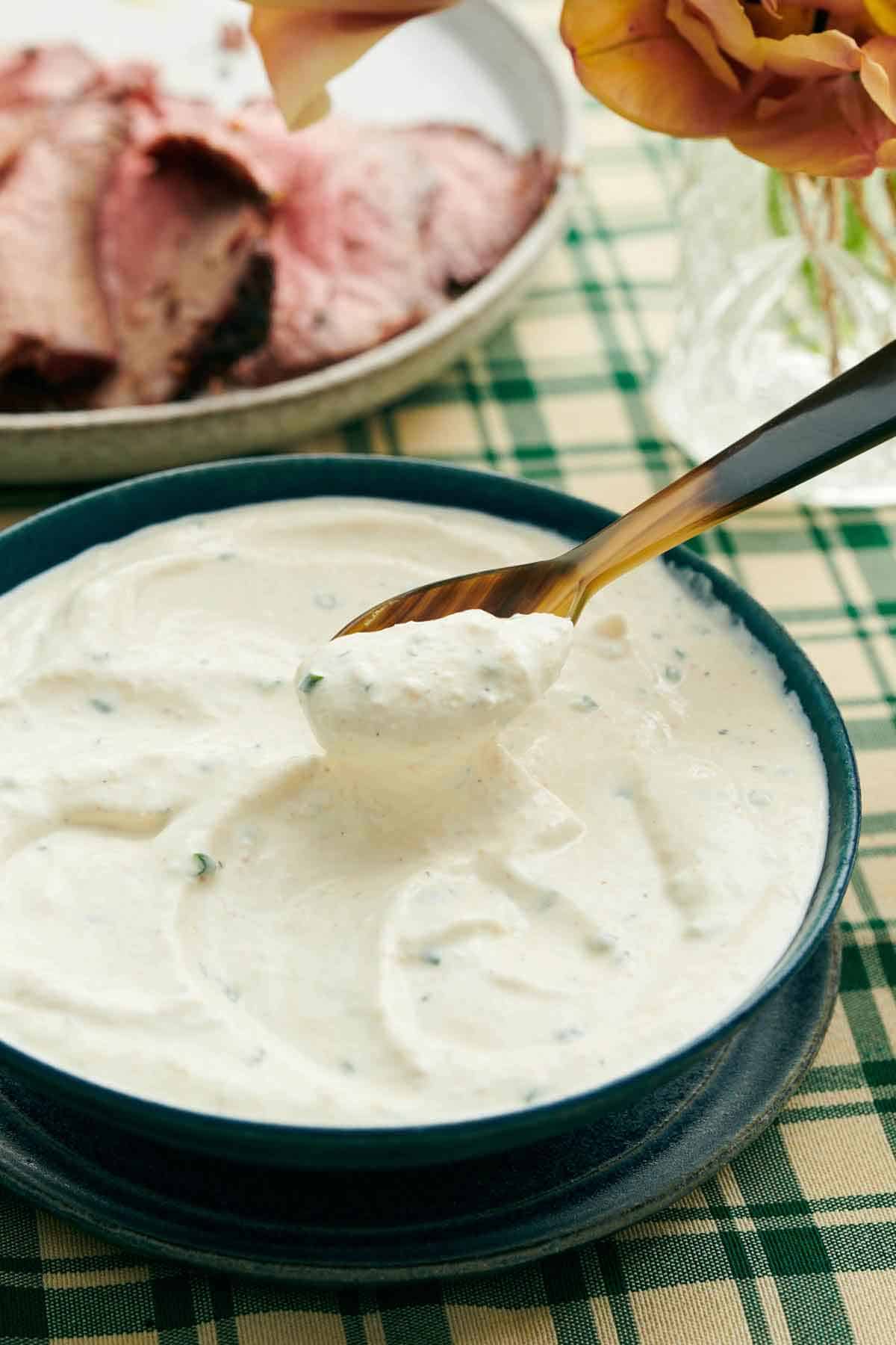 A spoonful of horseradish sauce lifted from a bowl. Flowers and sliced beef in the background.