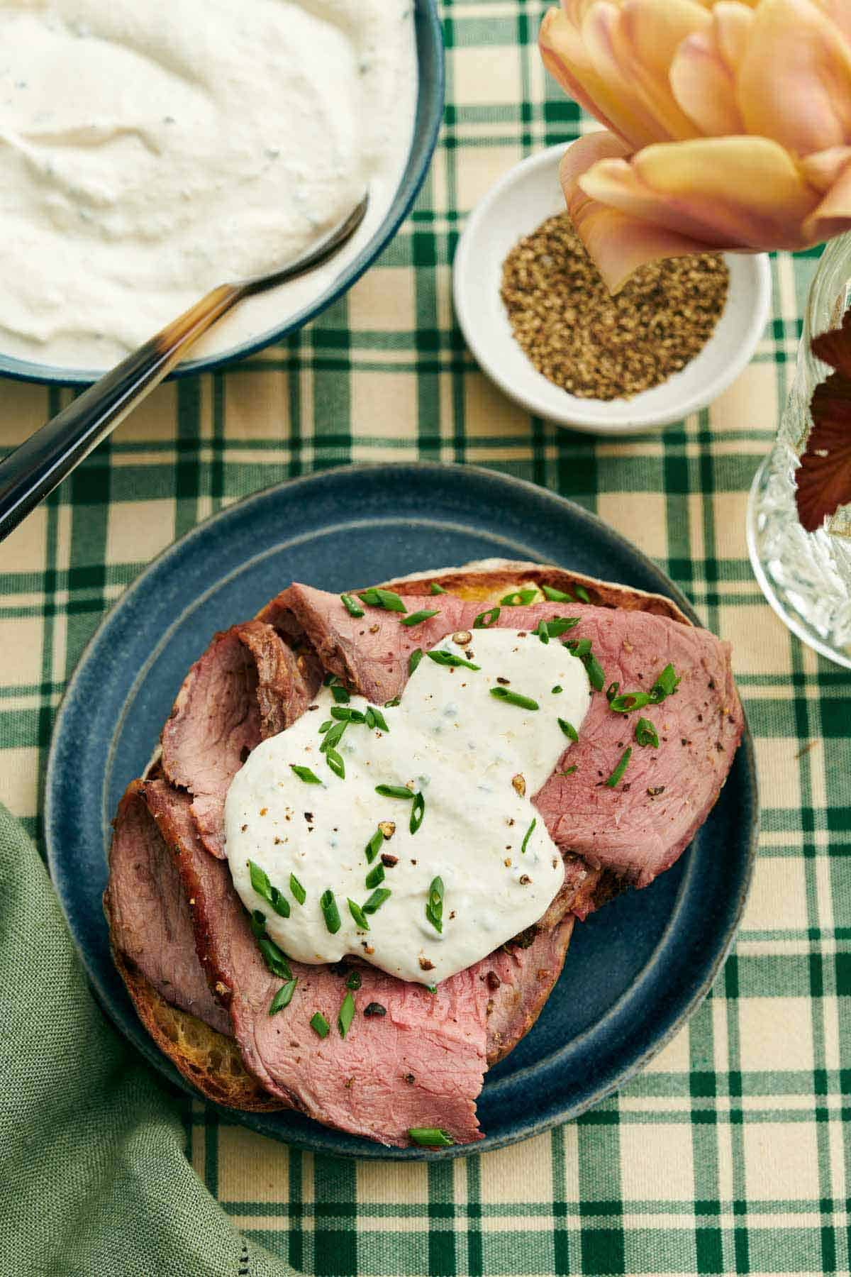 Overhead view of horseradish sauce over sliced beef topped with chopped chives. A bowl of more horseradish sauce on the side beside a bowl of pepper and flowers.