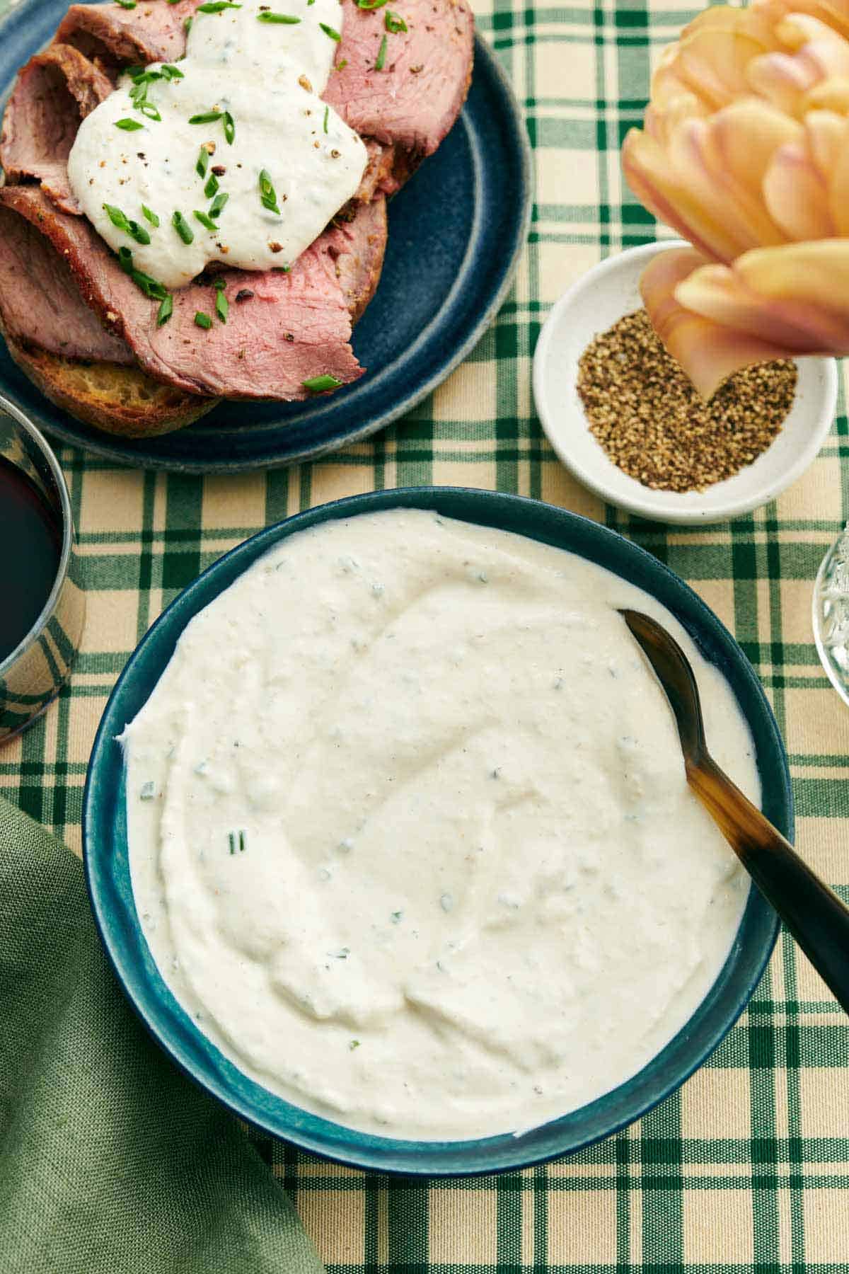 Overhead view of a bowl of homemade horseradish sauce with a spoon. A plate of sliced beef with horseradish sauce and chive garnish, a bowl of pepper, and flowers off to the side.