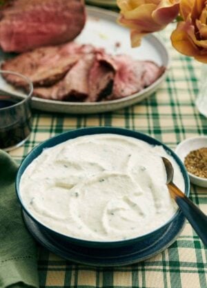 A bowl of horseradish sauce with a spoon. Sliced beef on a platter in the background with some flowers, bowl of pepper, and a drink.