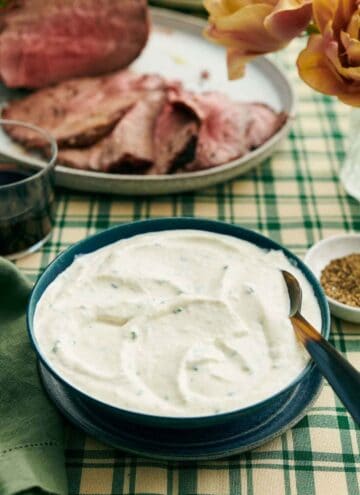 A bowl of horseradish sauce with a spoon. Sliced beef on a platter in the background with some flowers, bowl of pepper, and a drink.