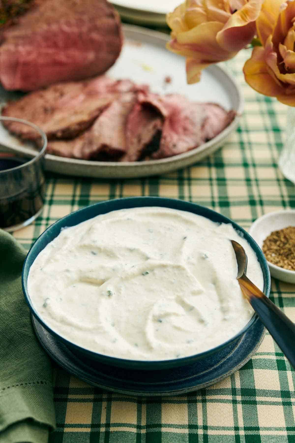 A bowl of horseradish sauce with a spoon. Sliced beef on a platter in the background with some flowers, bowl of pepper, and a drink.