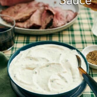 Pinterest graphic of a bowl of horseradish sauce with a spoon. Sliced beef on a platter in the background with some flowers and bowl of pepper.