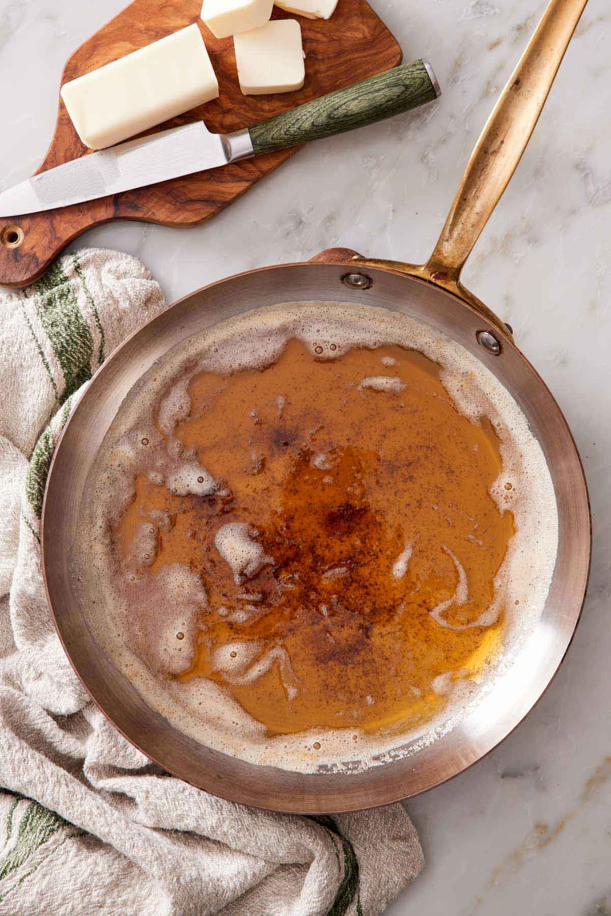 Overhead view of a skillet showing how to brown butter. A knife with sliced butter on the side on a wooden board.