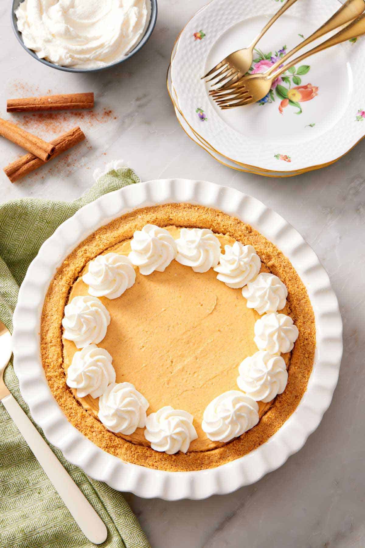 Overhead view of a pie dish with a no-bake pumpkin cheesecake topped with dollops of whipped cream. A stack of plates, forks, cinnamon sticks, and bowl of whipped cream on the side.