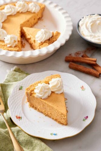 A slice of no-bake pumpkin cheesecake on a plate with a baking dish with the rest of the cheesecake in the background. Cinnamon sticks and a bowl of whipped cream on the side.