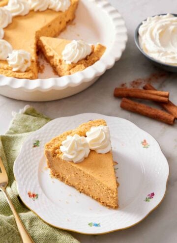 A slice of no-bake pumpkin cheesecake on a plate with a baking dish with the rest of the cheesecake in the background. Cinnamon sticks and a bowl of whipped cream on the side.