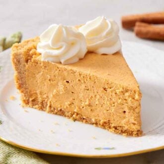 A plate with a slice of no-bake pumpkin cheesecake with two dollops of whipped cream on top. Cinnamon sticks in the background.