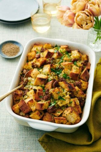 A baking dish of sausage stuffing with a spoon inside. Flowers, drinks, bowl of pepper, and plates in the background.