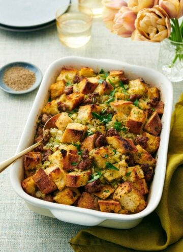 A baking dish of sausage stuffing with a spoon inside. Flowers, drinks, bowl of pepper, and plates in the background.