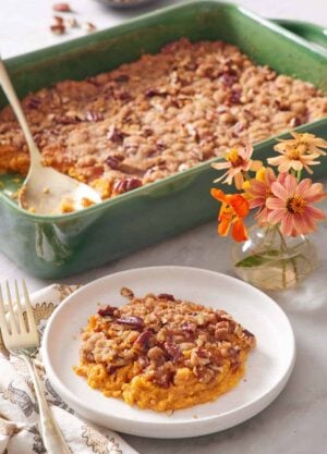 A plate with a serving of sweet potato casserole with flowers in the background along with the rest of the casserole with a spoon.