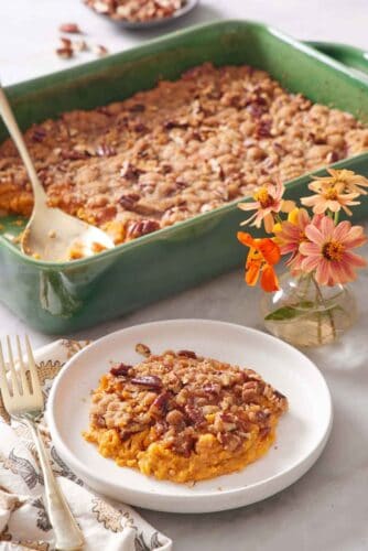 A plate with a serving of sweet potato casserole with flowers in the background along with the rest of the casserole with a spoon.