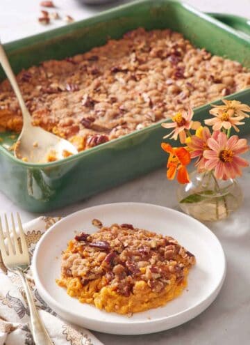 A plate with a serving of sweet potato casserole with flowers in the background along with the rest of the casserole with a spoon.