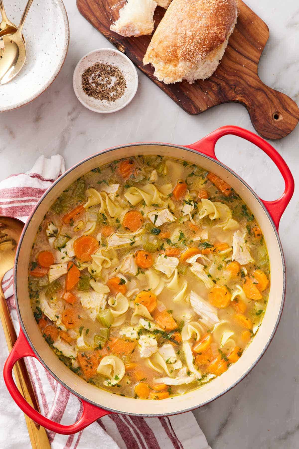 Overhead view of a pot of turkey noodle soup. Torn bread on a wooden serving board, bowl of pepper, and bowls with spoons off to the side.