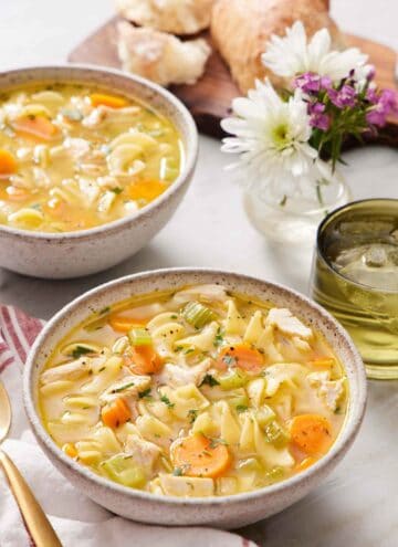 Two bowls of turkey noodle soup with a drink, flowers, and bread in the background.