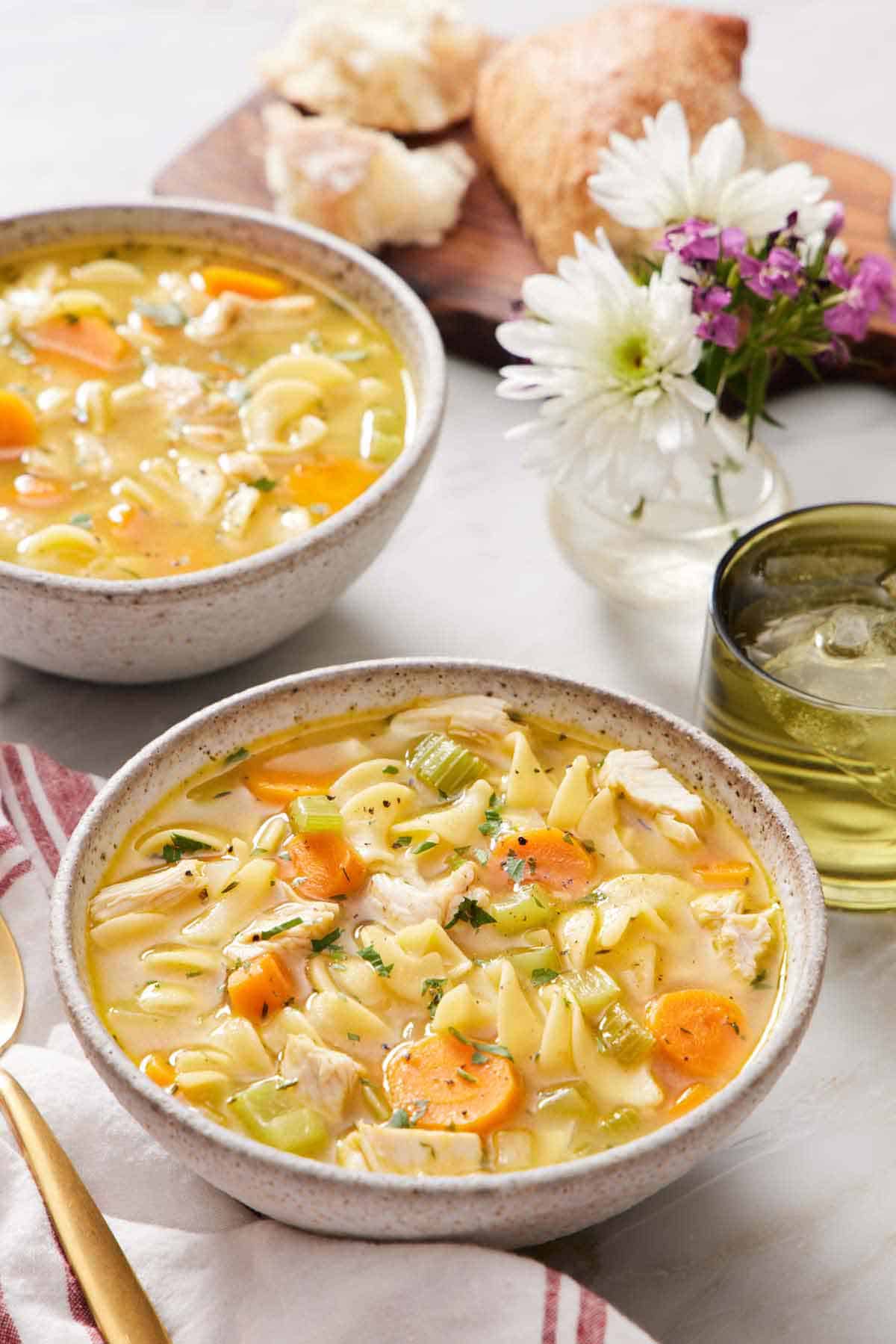 Two bowls of turkey noodle soup with a drink, flowers, and bread in the background.