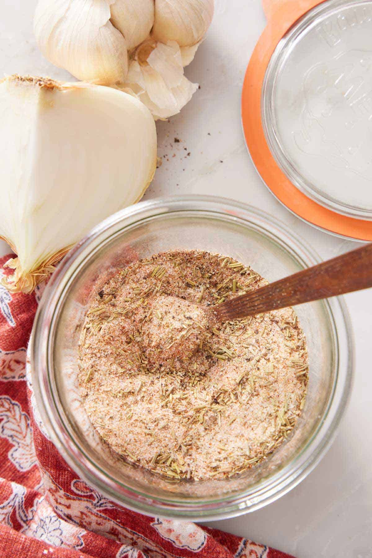 An overhead view of a jar of turkey seasoning with a measuring spoon inside. A cut onion, garlic, and jar lid on the side.