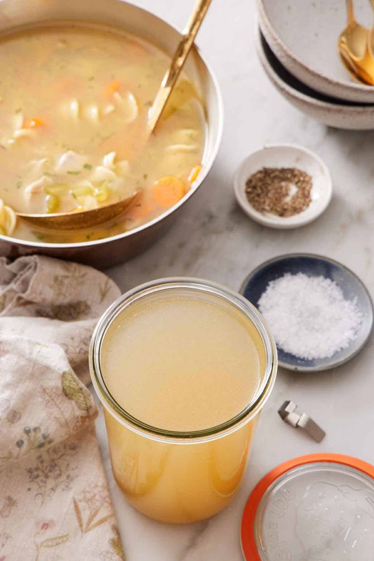 A jar of turkey stock with a pot of soup in the background along with a bowl of salt, pepper, and bowls.