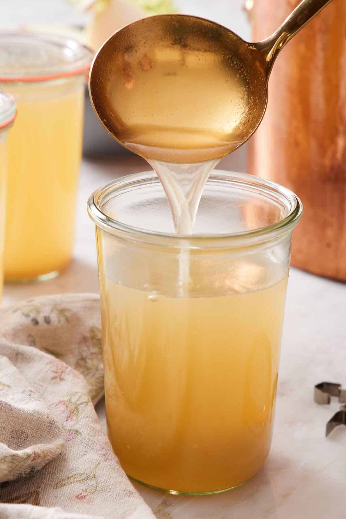 A ladle pouring turkey stock into a tall jar. Filled jars in the background along with a stock pot.