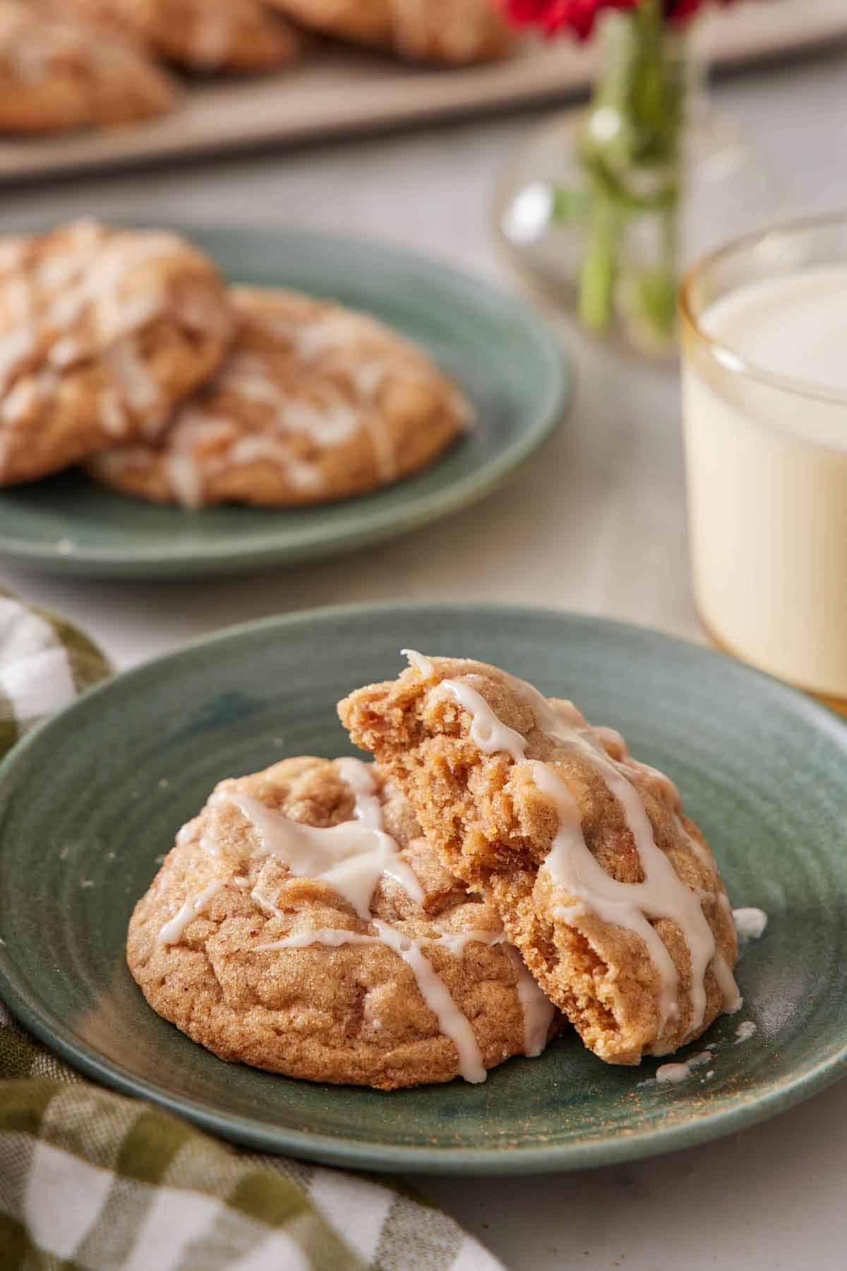 A plate with a brown butter wassail cookie with a half cookie leaning on top. A cup of milk and plate of more cookies in the background.