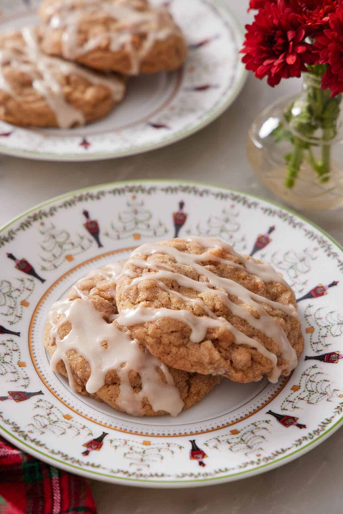 A plate with two brown butter wassail cookies with icing drizzled on top. Two more on a plate in the background along with some flowers.