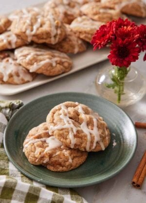 A plate with two brown butter wassail cookies with a platter more in the background along with some red flowers in a small vase.