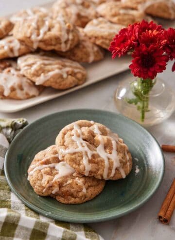 A plate with two brown butter wassail cookies with a platter more in the background along with some red flowers in a small vase.